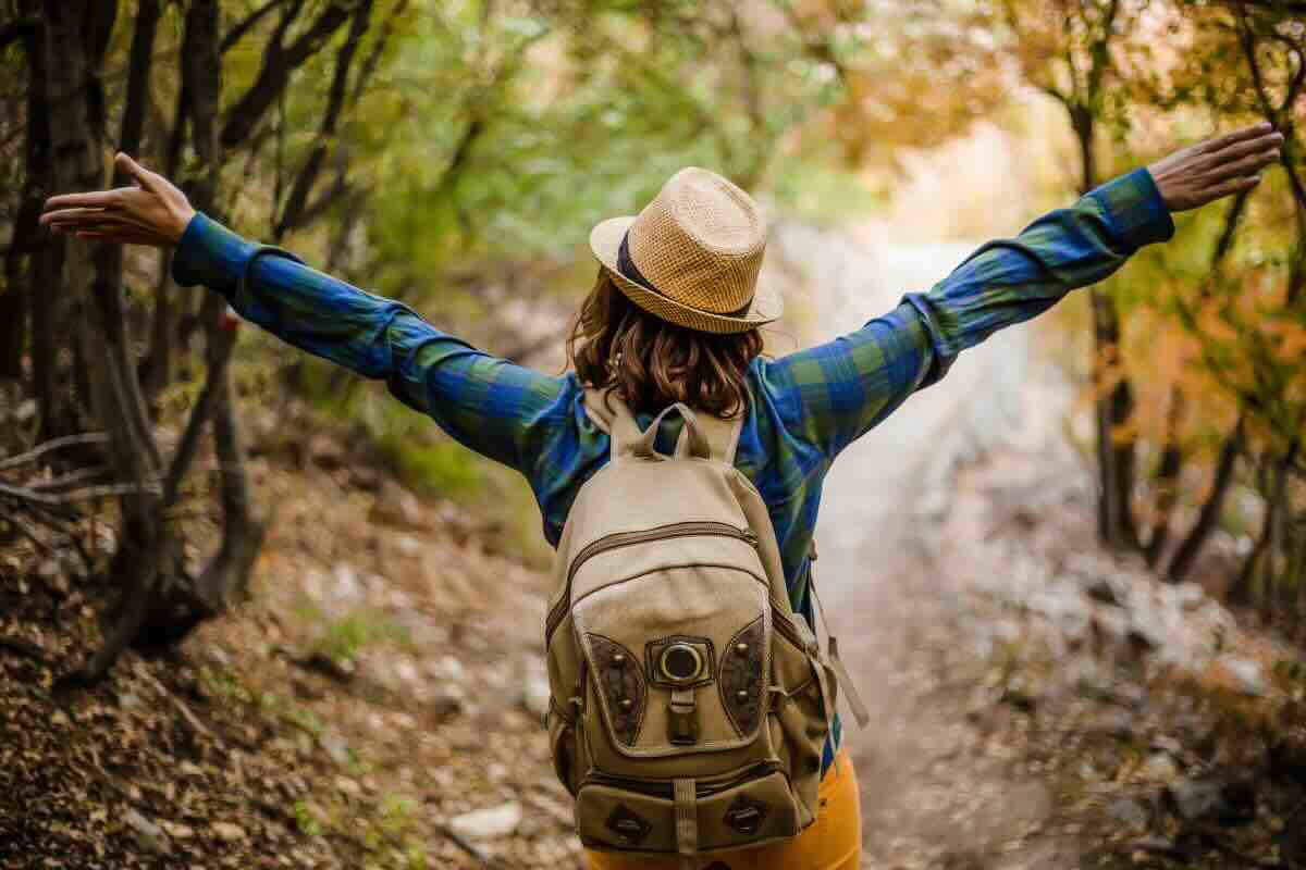 Donna di spalle con braccia spalancate, cappello e zaino, nella natura